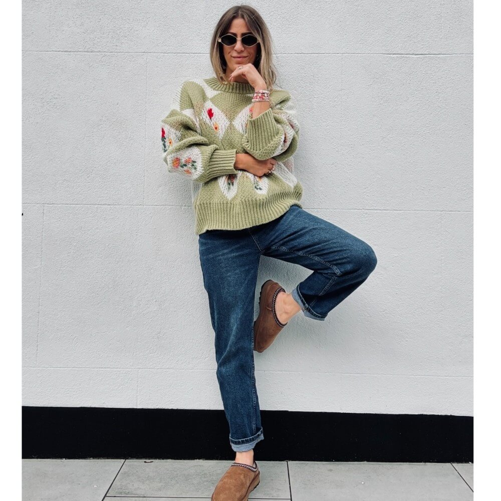 Woman wearing a green sweater with floral patterns and blue jeans, leaning against a light gray wall. EMU Sharky Scuff Chestnut