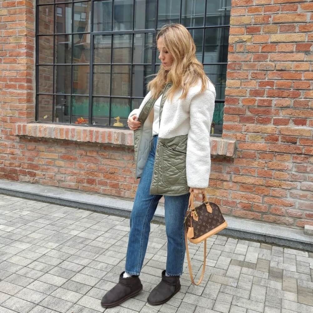 Woman standing outdoors against a brick wall, wearing a white jacket blue jeans And EMU Stinger Micro Chocolate