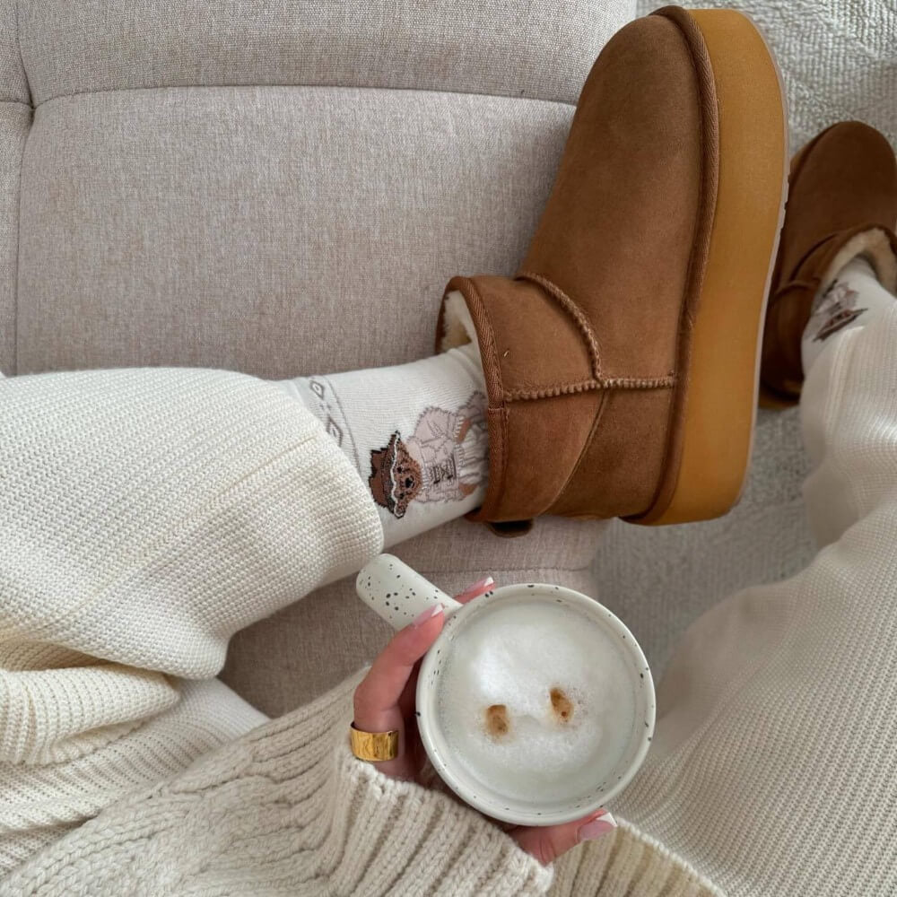Person wearing brown slippers and holding a cup, sitting on a beige couch. EMU Stinger Micro Flatform Chestnut
