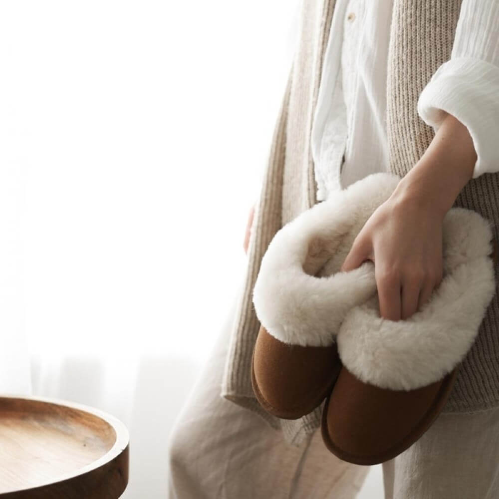 Person wearing brown slippers with white fuzzy trim, holding one slipper. EMU Australia Jolie Slipper Chestnut
