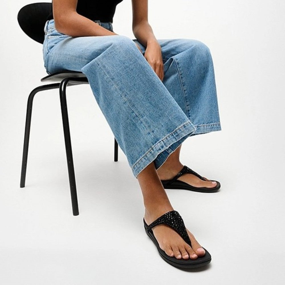 Person wearing black fitflop sandals and blue jeans sitting on a chair.