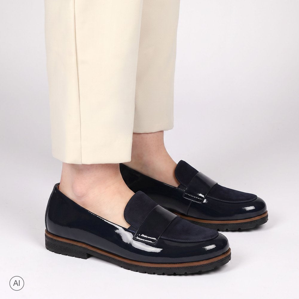 A pair of Gabor glossy navy patent leather penny loafers worn with cream trousers, shown from the calves down against a clean white studio background.