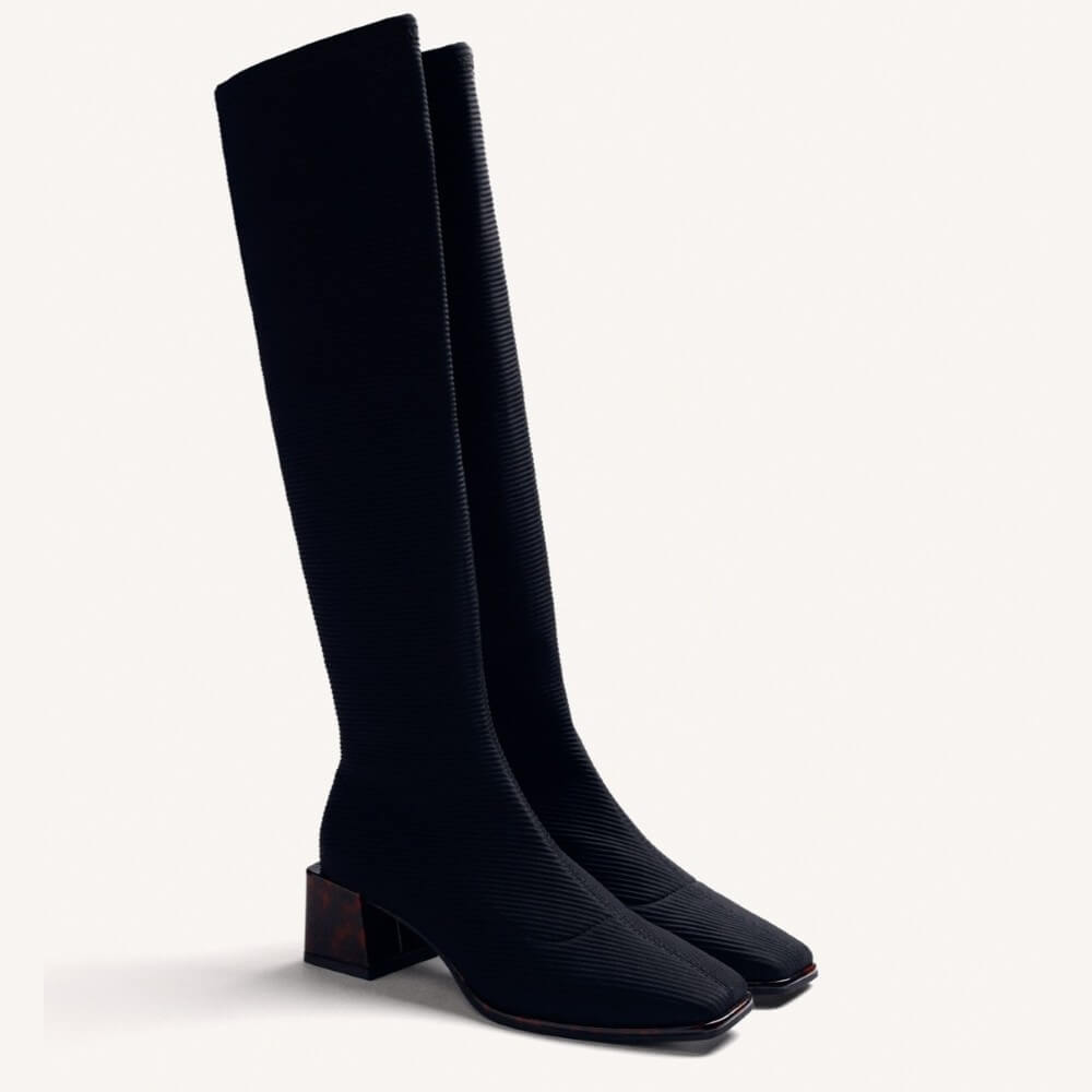 A pair of black knee-high boots with a textured, ribbed fabric upper and a low block heel, shown against a plain off-white background.