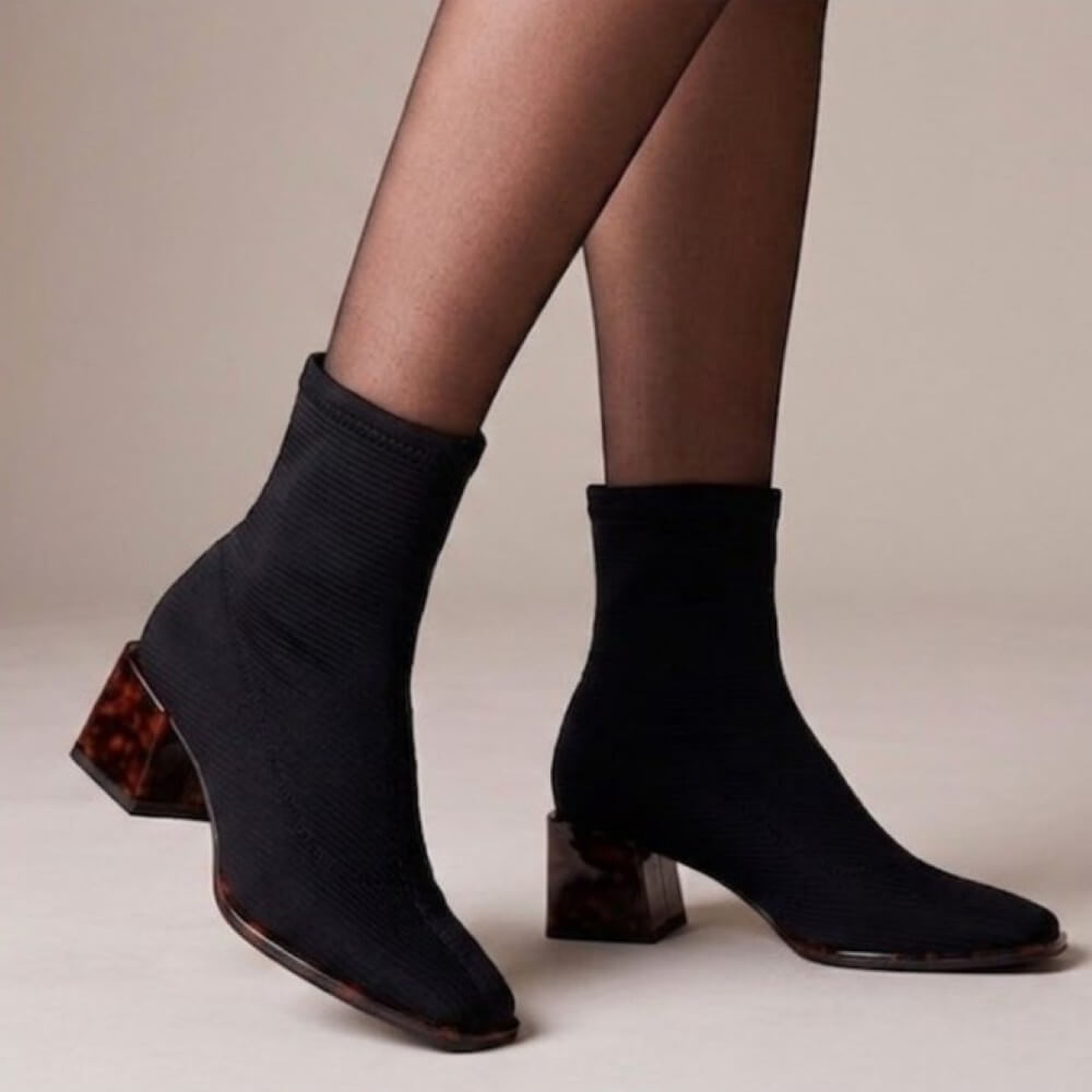 Gioseppo Black knit ankle boots with chunky square block heels and tortoiseshell trim, shown on a model's feet against a neutral beige background.