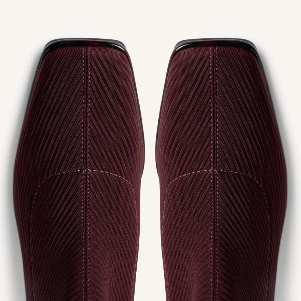 Close-up of a pair of burgundy textured boots with diagonal ribbed fabric and contrast stitching, shown against a light off-white background.