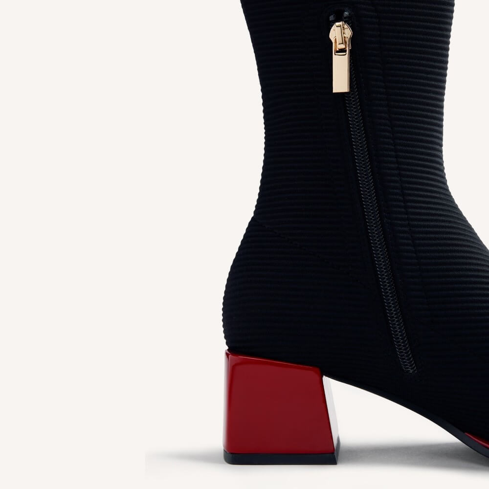 Close-up of a Gioseppo black ribbed knit ankle boot with a glossy red block heel and a gold zipper pull, shown on a light off-white background.