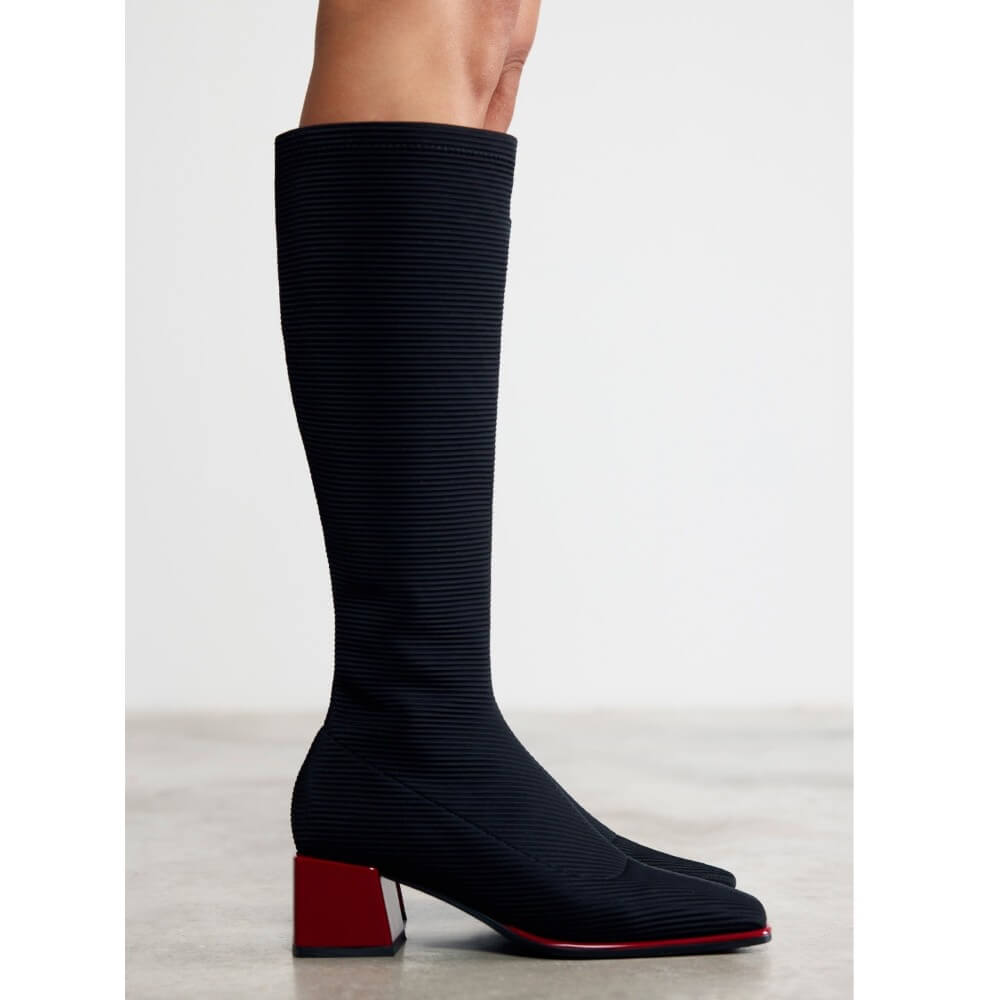 Gioseppo knee-high ribbed knit boots in black with a bright red block heel and red outsole, shown on a model against a light gray studio background.
