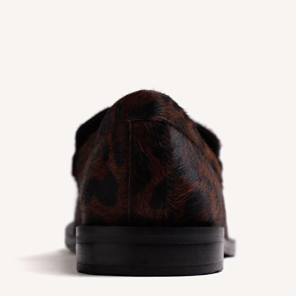 A close-up rear view of a brown and black leopard-print loafer with a thick black sole, set against a plain off-white background.