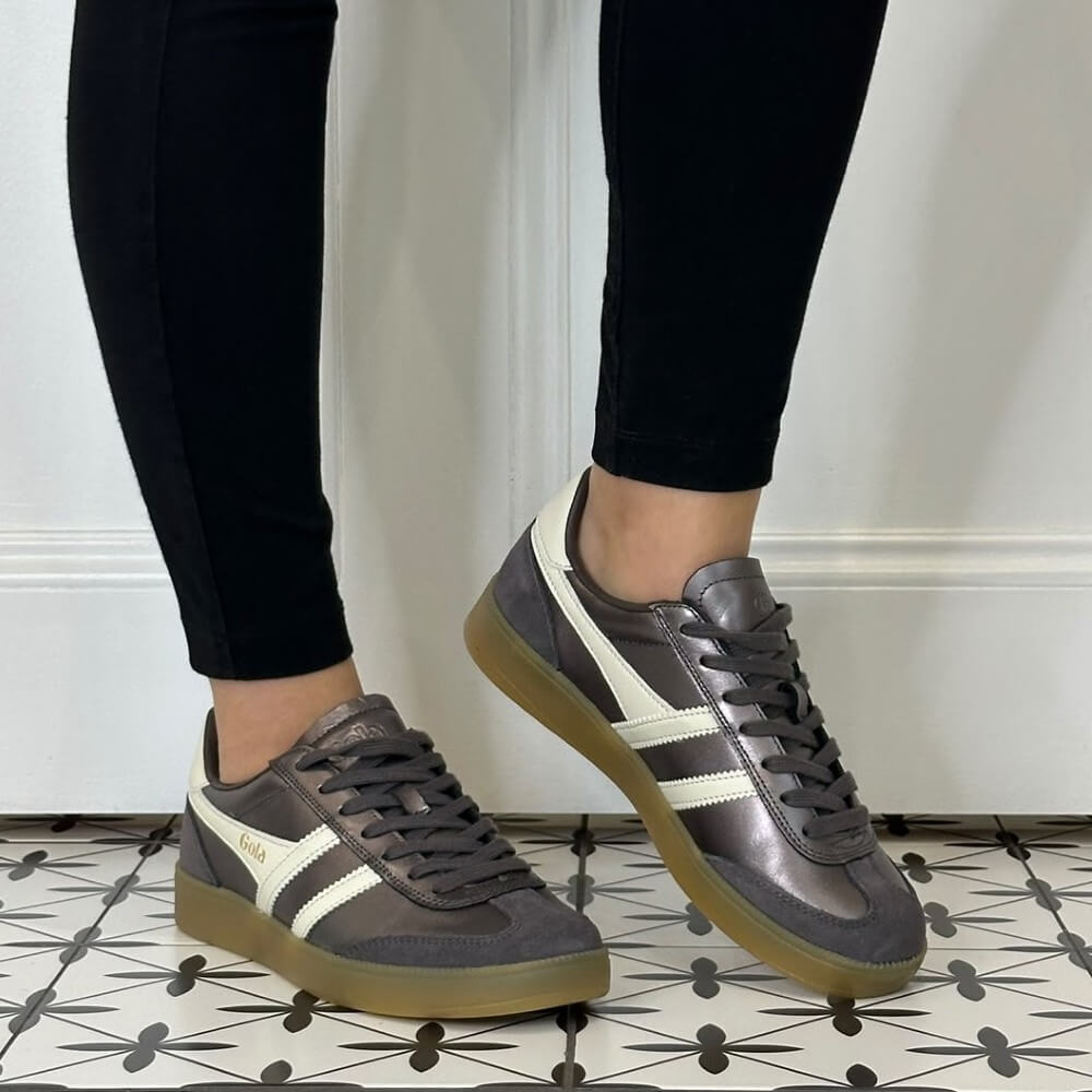 A person wearing black leggings and Gola grey low-top sneakers with white side stripes and gum soles, standing on a patterned tile floor in front of a white panelled wall.