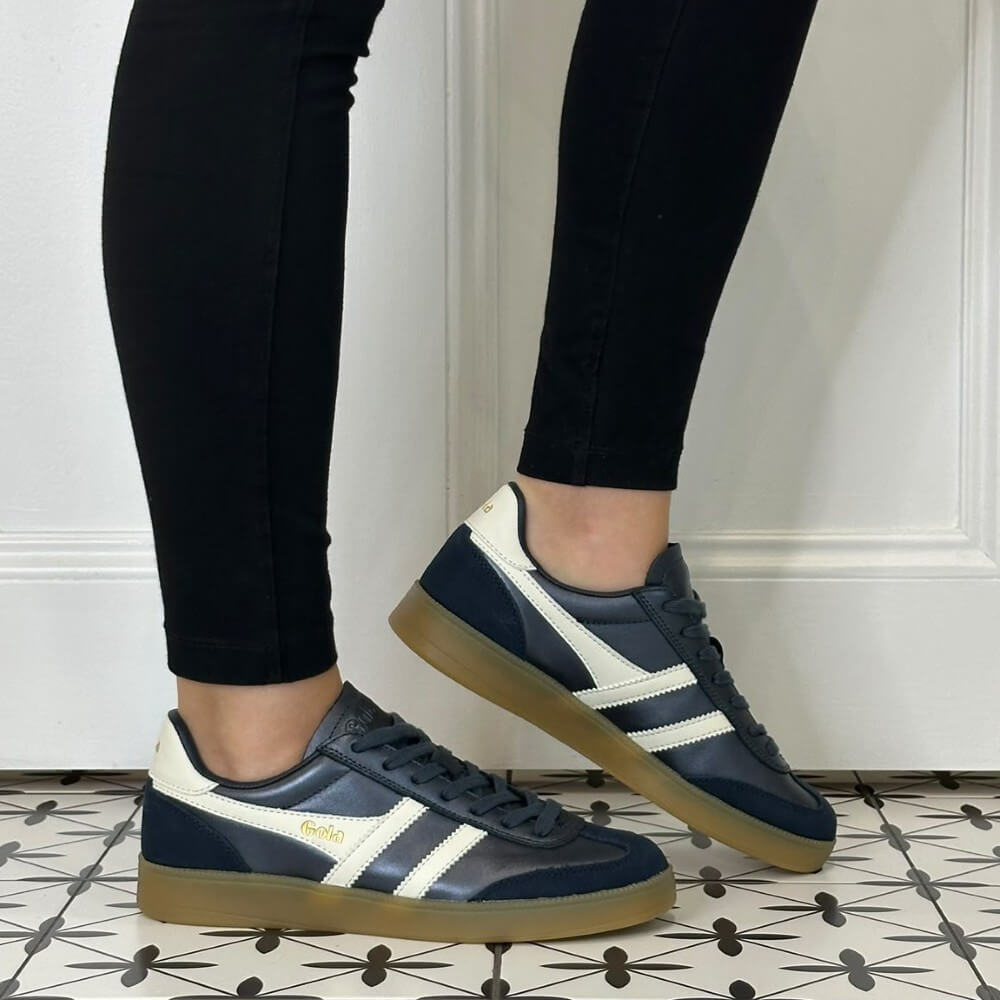 A person wearing Gola navy blue and off-white sneakers with gum soles, paired with black leggings, standing on a patterned tile floor in front of a white panelled wall.
