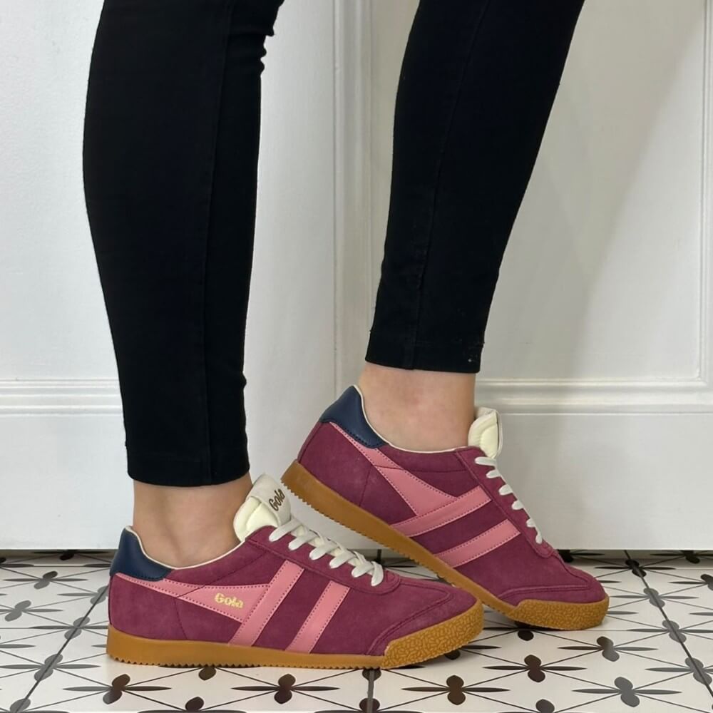 A person wearing black skinny jeans and Gola pink and burgundy suede Gazelle sneakers with a gum sole, standing on a patterned tile floor against a white panelled wall.