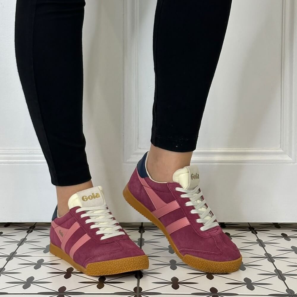 A person wearing black skinny jeans and Gola pink and burgundy suede Gazelle sneakers with a gum sole, standing on a patterned tile floor against a white panelled wall.