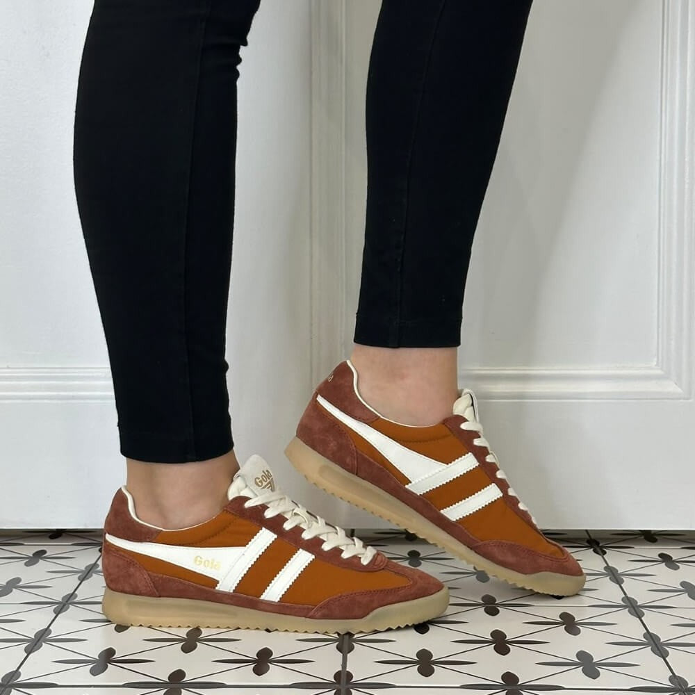  A person wearing Gola brown suede and canvas low-top sneakers with white stripes and white laces, paired with black leggings, standing on a black and white patterned tile floor against a white wall.