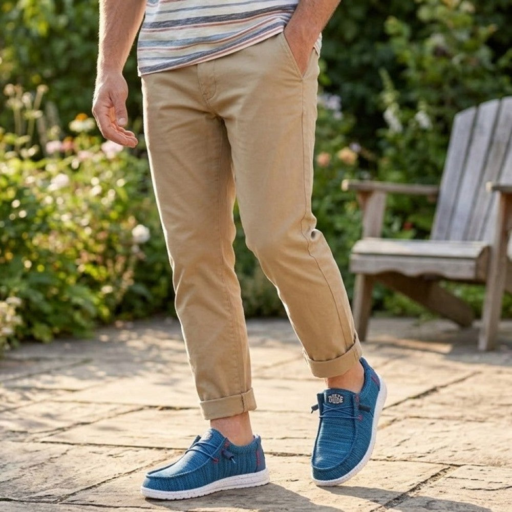 Person wearing HeyDude blue shoes and beige pants standing on a paved area with greenery in the background