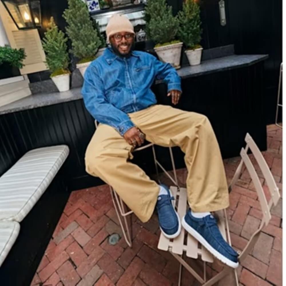 Man sitting on a stool wearing a blue jacket, beige pants, and HeyDude blue shoes in an outdoor setting.