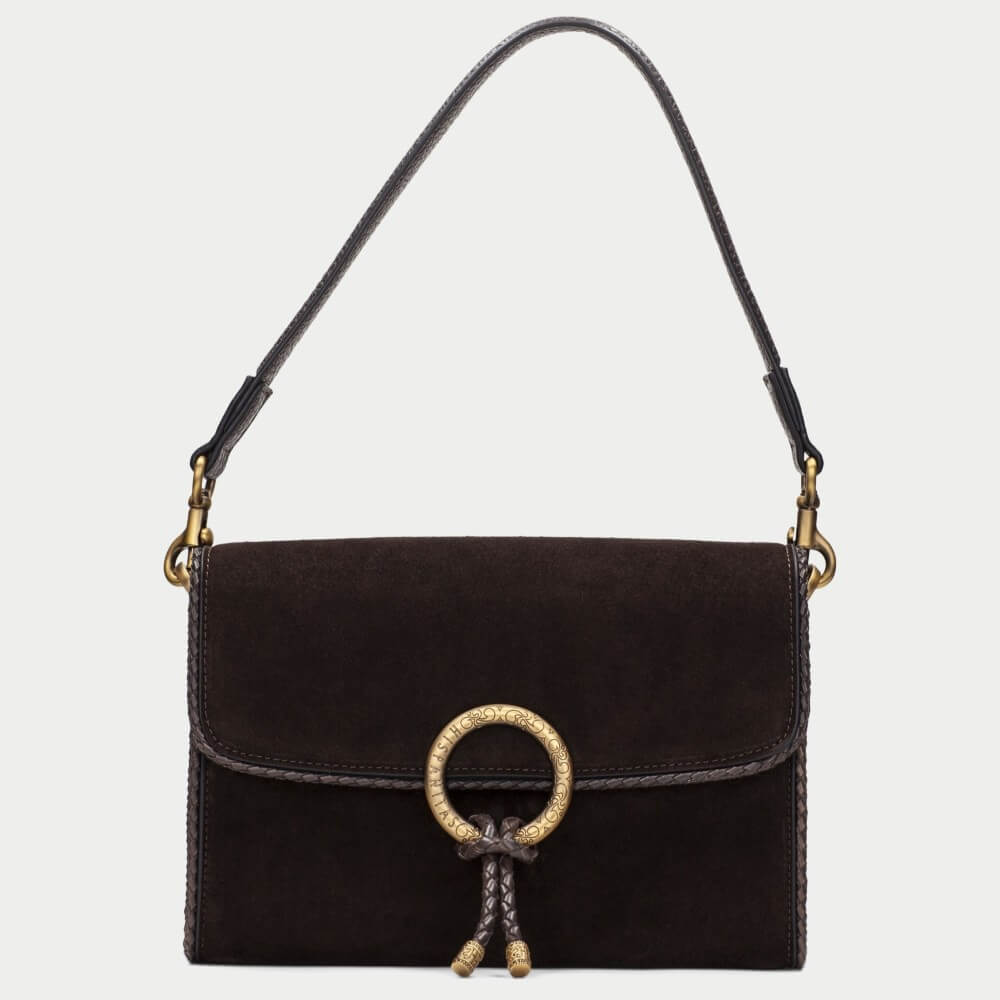 A Hispanitas dark brown suede shoulder bag with a gold-tone circular ring clasp and braided tassel detail, shown against a light gray background.