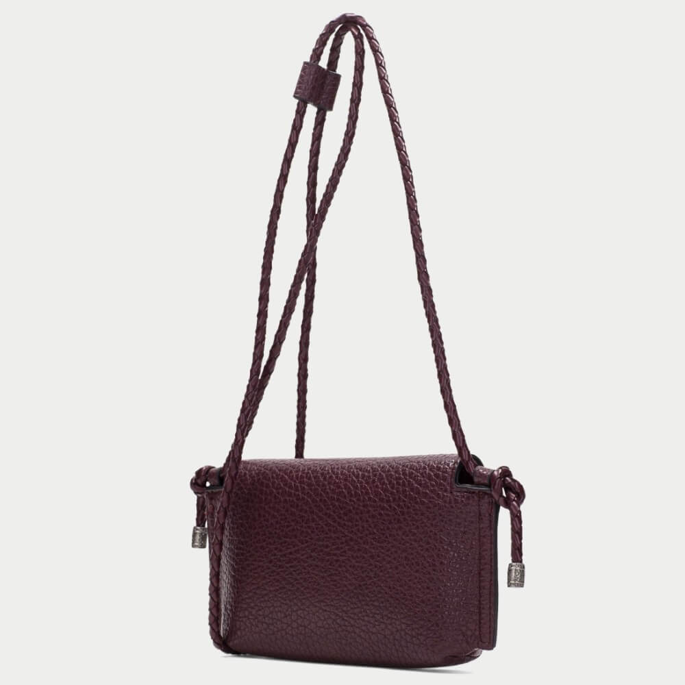A Hispanitas burgundy pebbled PU leather clutch bag with a flap closure and braided strap details,   set against a plain light grey background.
