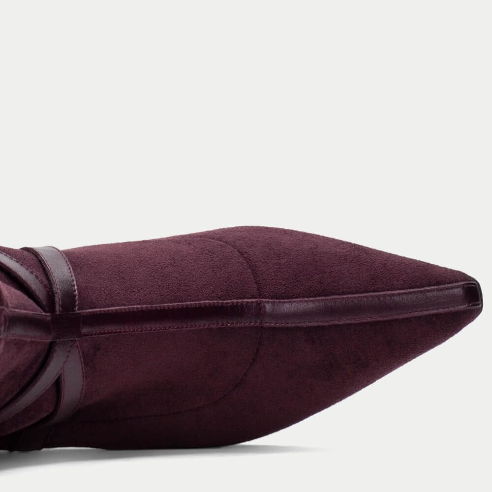 Close-up side view of a pointed-toe boot in deep burgundy suede with dark brown leather trim and straps, set against a plain light gray background.