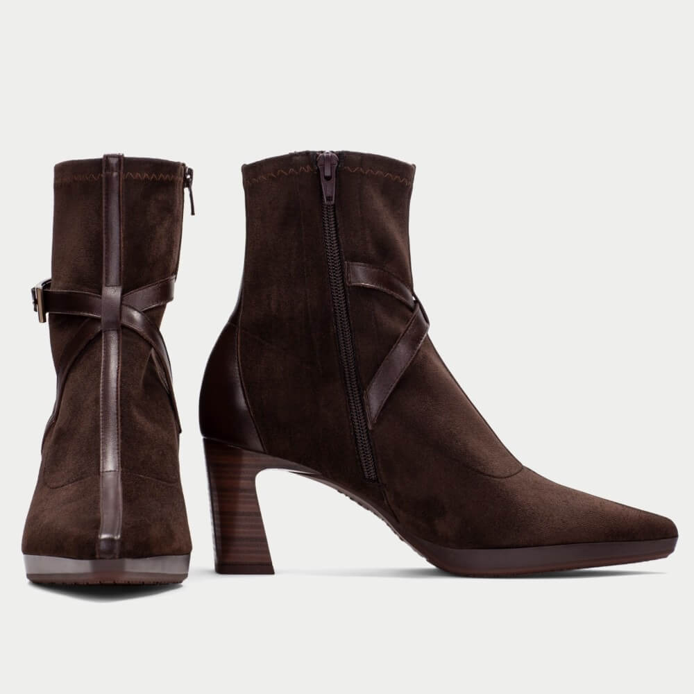 A pair of Hispanitas dark brown suede ankle boots with pointed toes, block heels, and decorative side straps, shown from the front and side on a light Grey background.