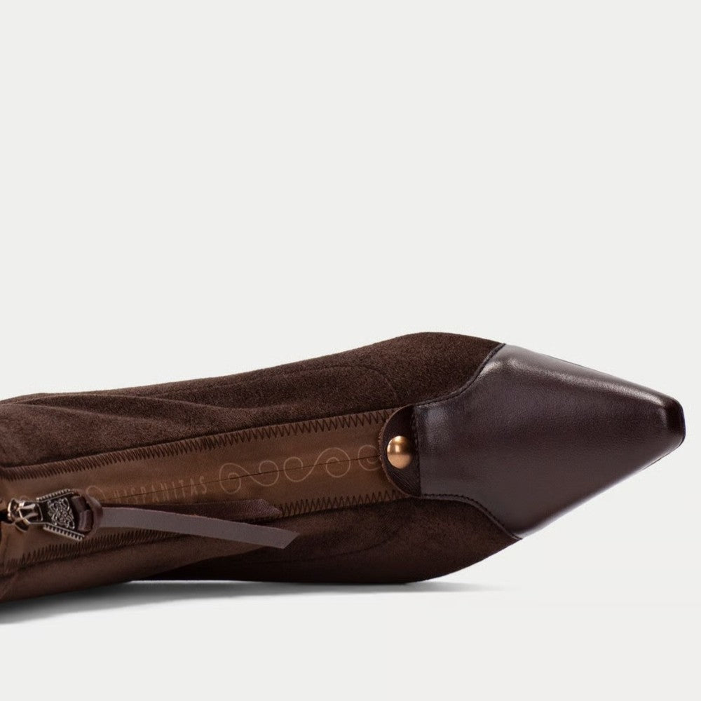 A close-up side view of a dark brown pointed-toe boot or shoe, showing a suede-like upper, smooth leather toe cap, zipper detail, and a small metallic snap accent against a light gray background.