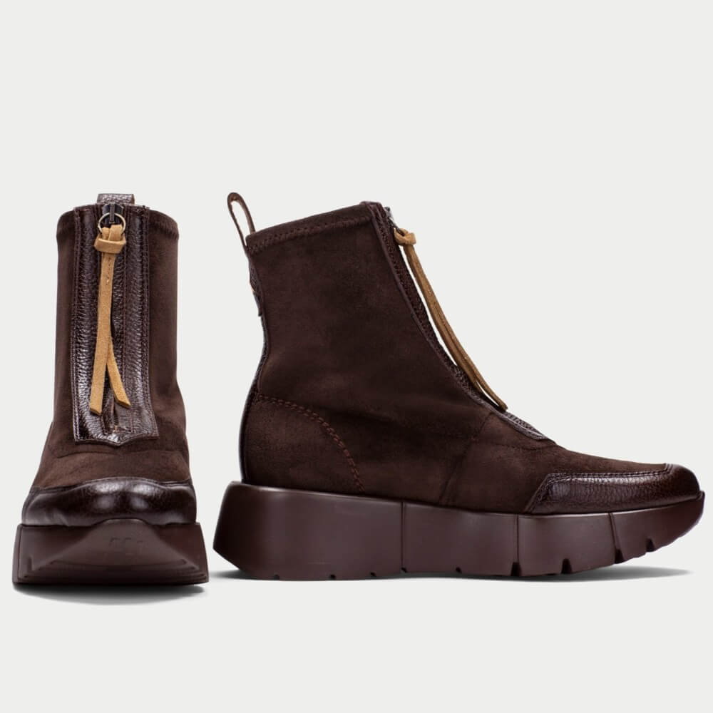 A pair of Hispanitas dark brown ankle boots with a chunky sole, featuring a side zipper with a leather pull tab and a front slip-on panel, shown from both front and side profiles against a light grey background.