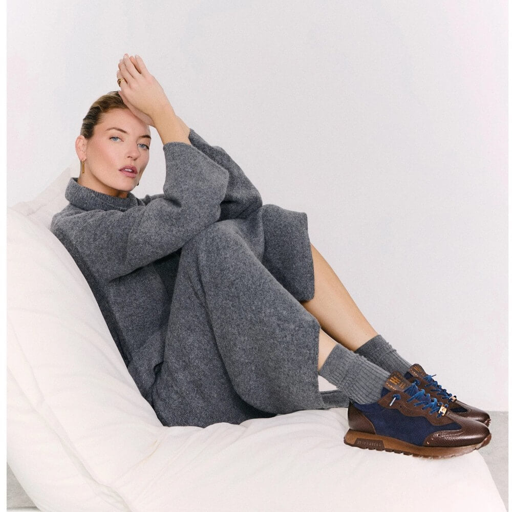 A woman reclines on a white beanbag chair wearing a loose, oversized grey knit sweater and matching grey knit pants, paired with Hispanitas brown and navy sneakers and grey socks.