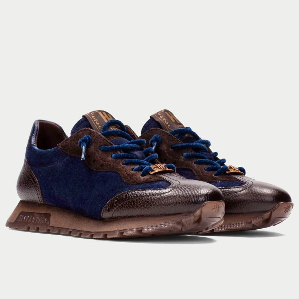 A pair of low-top sneakers by Hispanitas, featuring dark brown textured leather and navy blue suede panels, with matching navy laces and a brown sole, set against a light grey background.