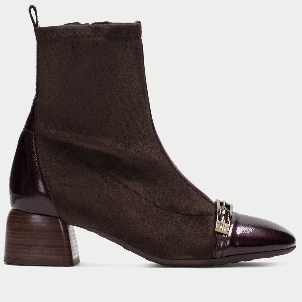 A Hispanitas dark brown ankle boot with a mix of matte suede and glossy patent leather panels, featuring a stacked block heel and a decorative gold-tone metal buckle on the toe.