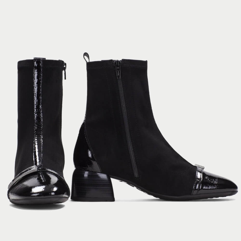 A pair of Hispanitas black ankle boots with a mix of matte suede and glossy patent leather textures, featuring a low block heel and a decorative silver-tone buckle detail on the toe cap, shown against a light gray background.