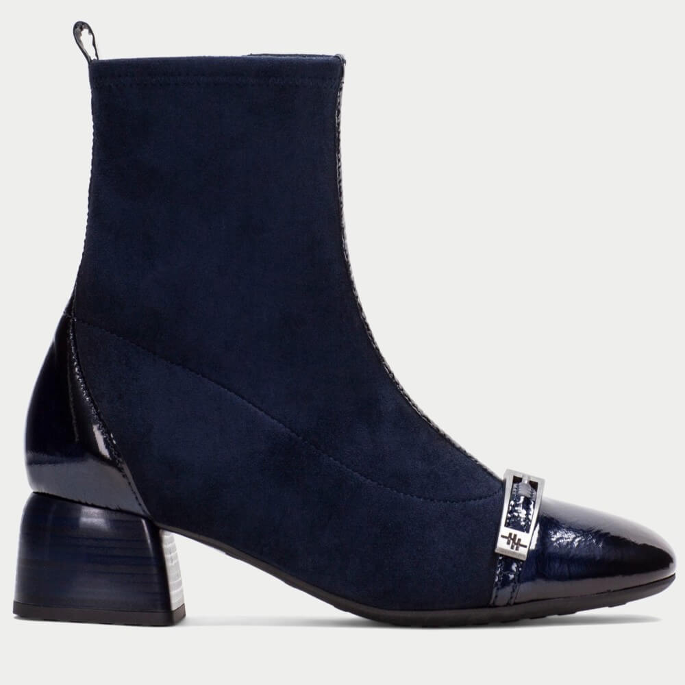 A Hispanitas navy blue ankle boot with a mix of suede and glossy patent leather panels, featuring a block heel and a decorative silver-tone metal buckle on the toe cap, shown against a light gray background.
