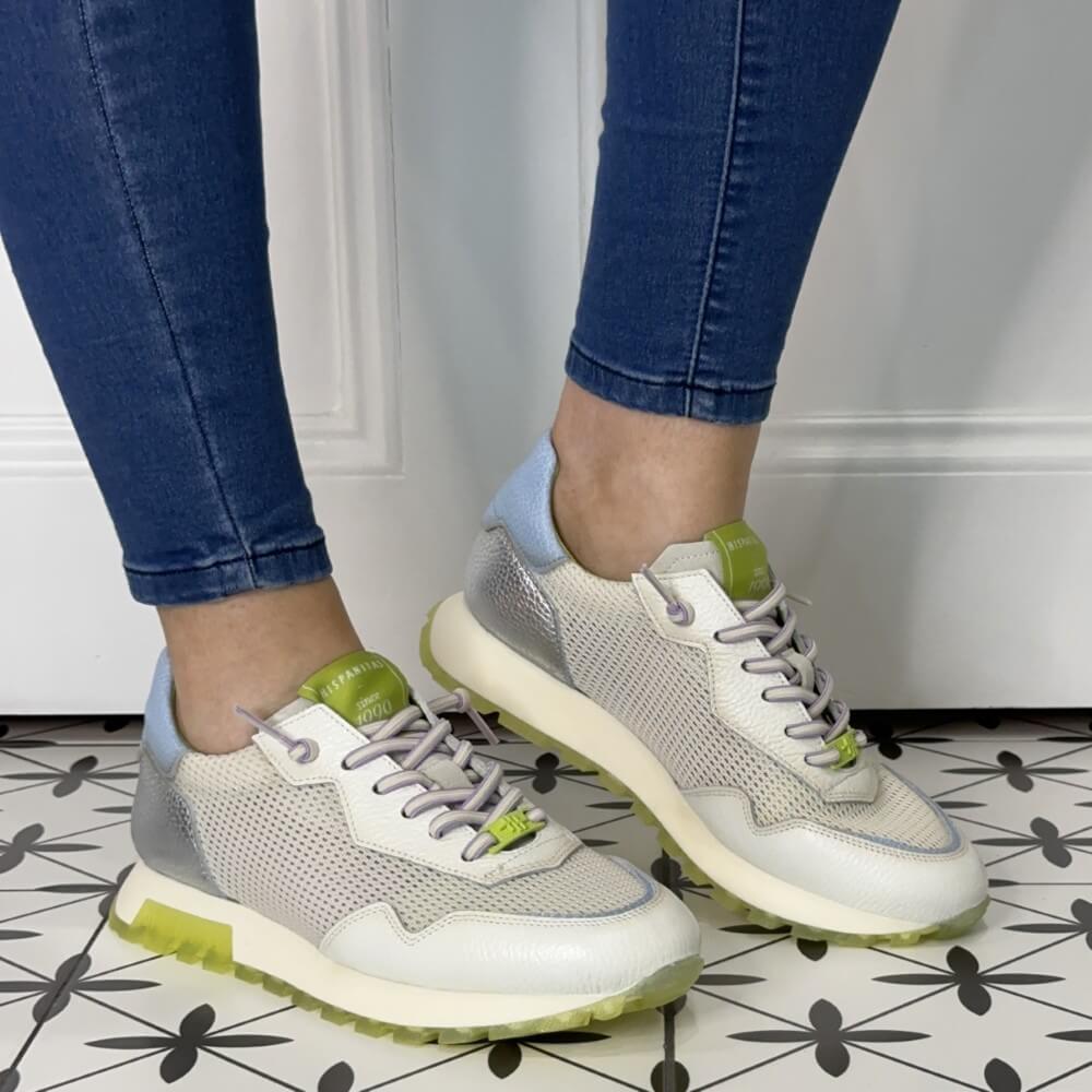 off white sneakers with green accents worn with blue jeans on a patterned floor.