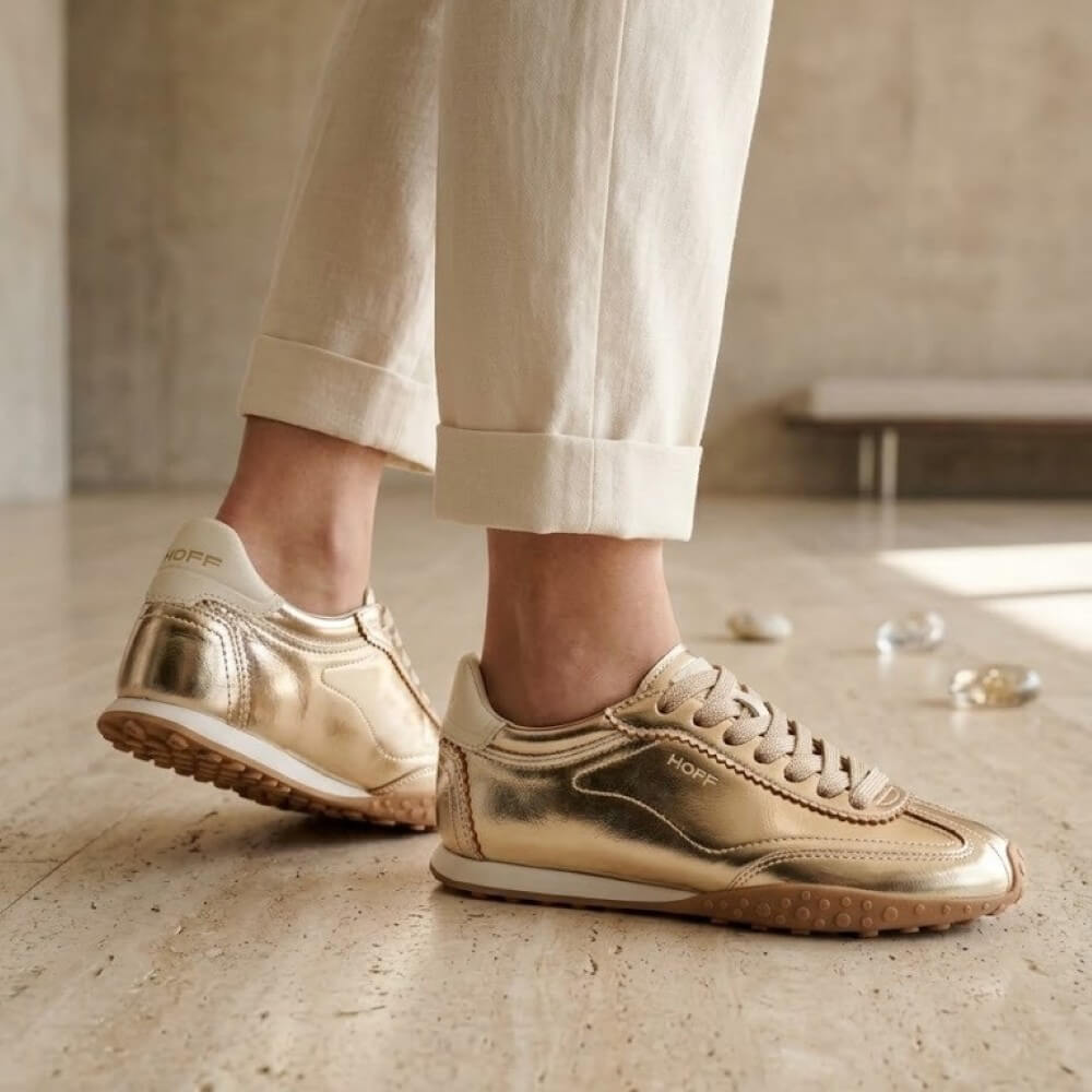 Hoff Metallic gold low-top sneakers worn with off-white cuffed trousers, shown on a person in a softly lit indoor setting with a stone floor.