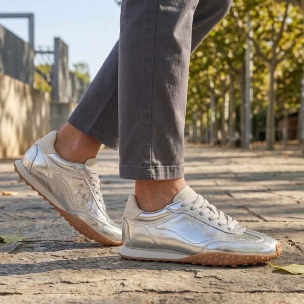 A person wearing Hoff shiny metallic silver sneakers with grey pants, standing on a stone pathway outdoors with trees in the background.