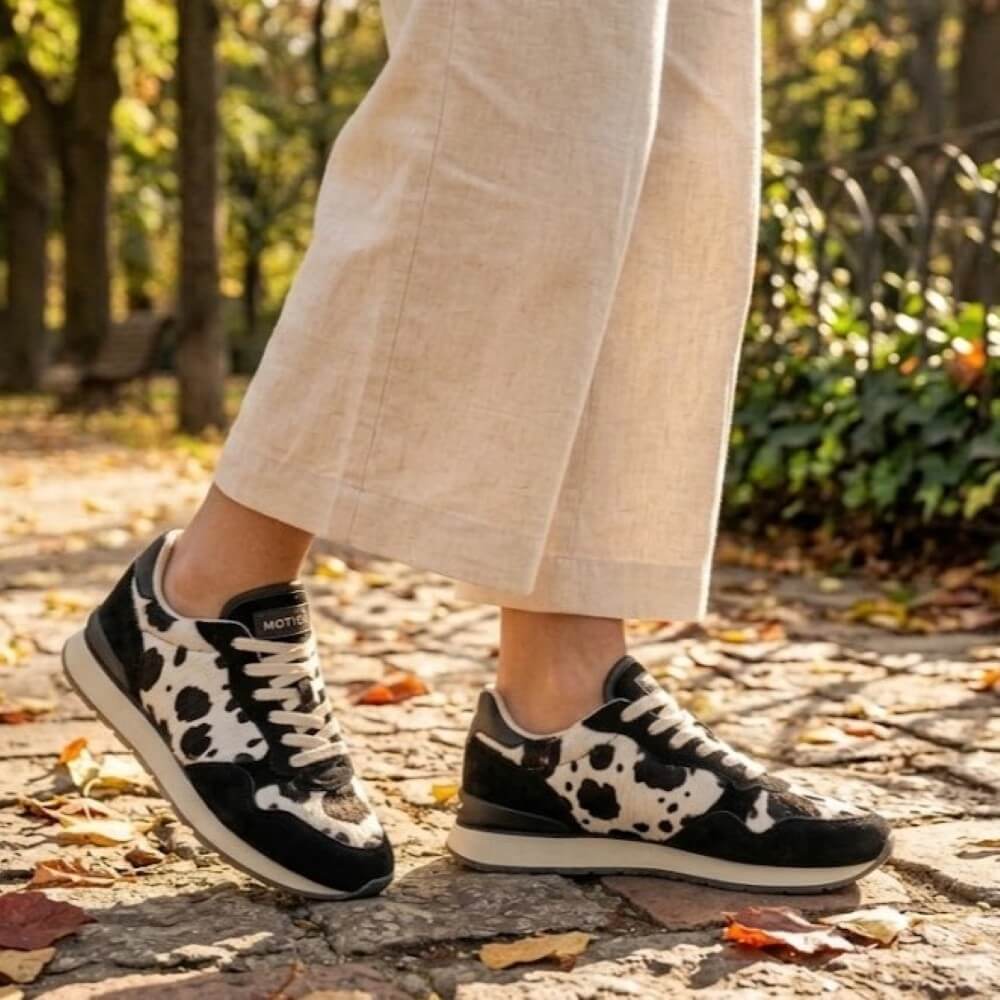 A person wearing Hoff black and white cow-print sneakers with beige cropped pants, standing on a stone path in a park with autumn leaves and trees in the background.