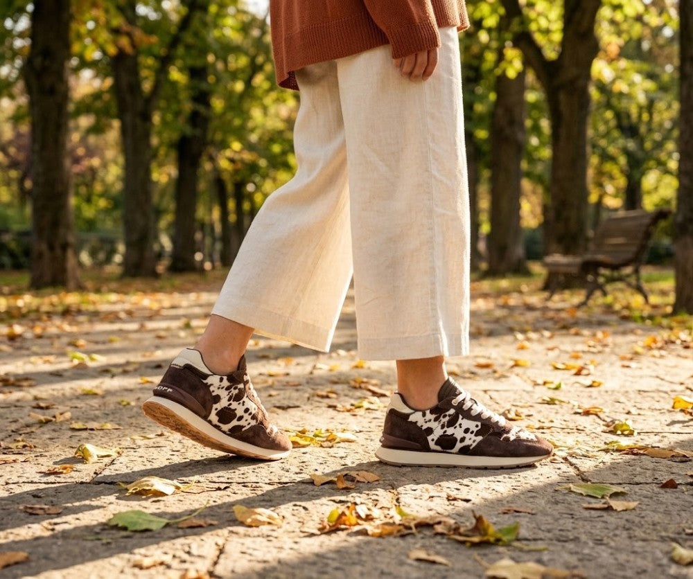 A person wearing off-white wide-leg trousers and Hoff brown low-top sneakers with a white cow-print pattern, standing on a leaf-strewn path in a sunny autumn park.