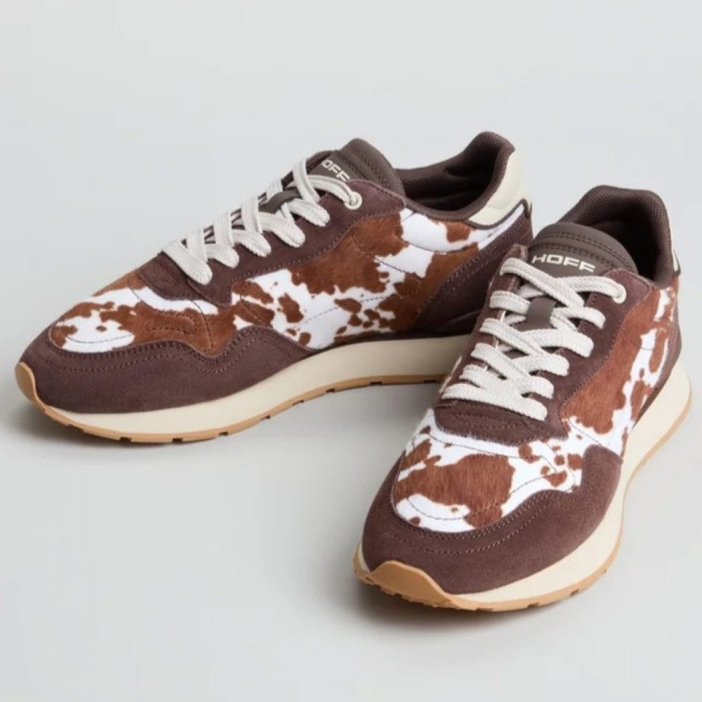 A pair of Hoff low-top sneakers with a brown and white cow-print pattern on the toe boxes and sides, dark brown suede overlays, white laces, and cream midsoles, shown on a light grey background.