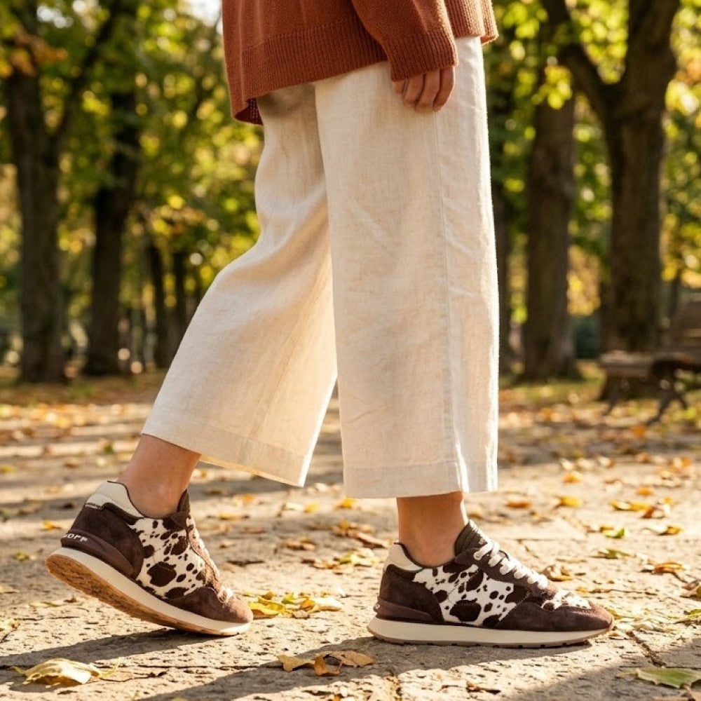 A person walking outdoors in a park wearingHoff brown and white cow-print sneakers, cream wide-leg cropped pants, and a rust-coloured knit sweater. The background shows blurred trees and fallen leaves.