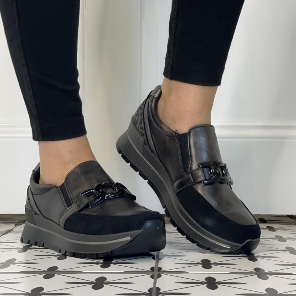 A person wearing Igi & Co  Grey slip-on platform sneakers with a decorative buckle detail, paired with black pants, standing on a patterned tile floor.