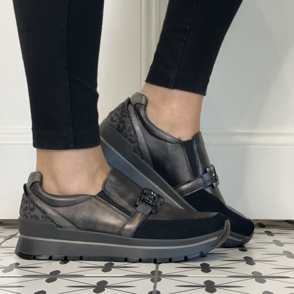 A person wearing Igi & Co  Grey slip-on platform sneakers with a decorative buckle detail, paired with black pants, standing on a patterned tile floor.