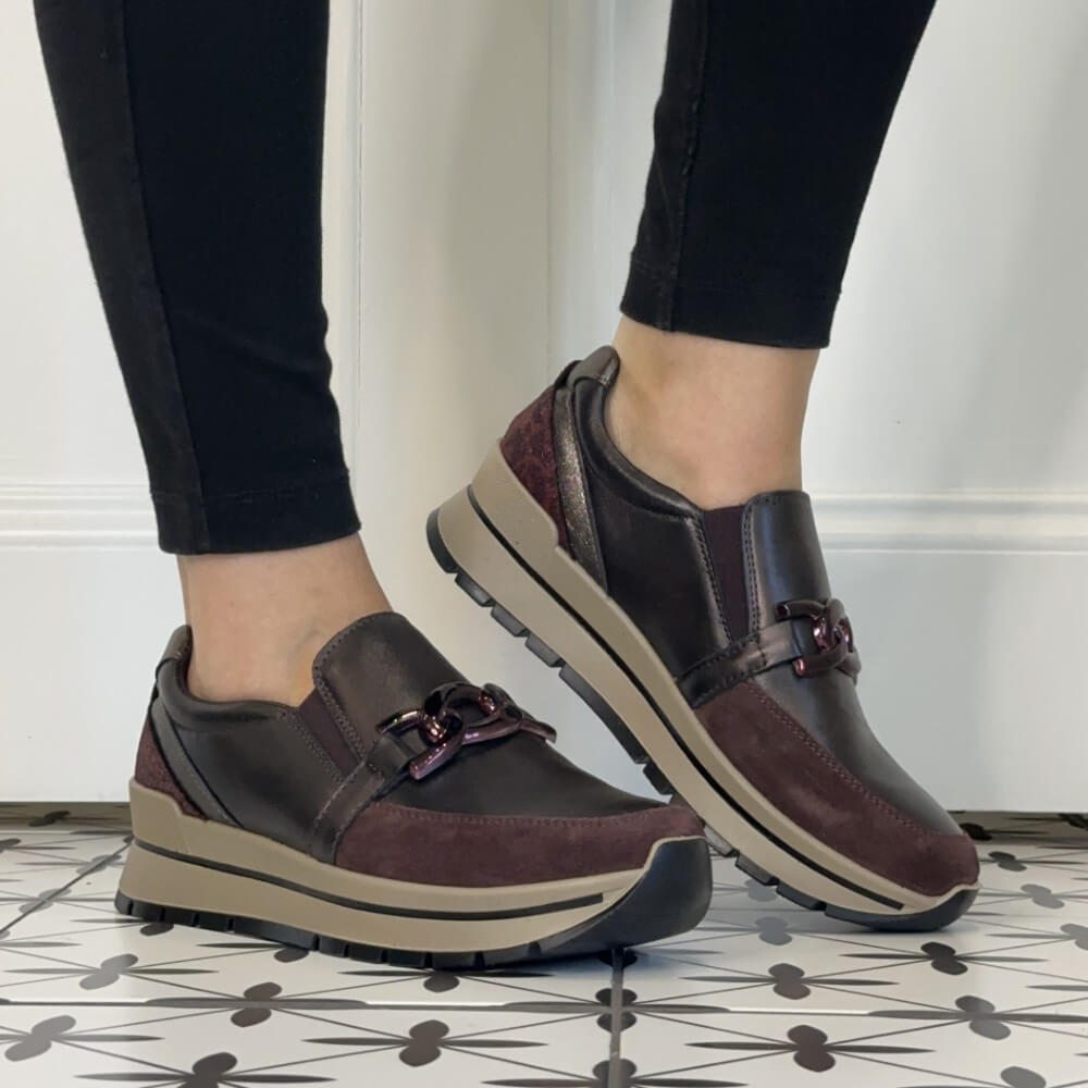 A person wearing Igi & Co   burgundy slip-on platform sneakers with a decorative buckle detail, paired with black pants, standing on a patterned tile floor.
