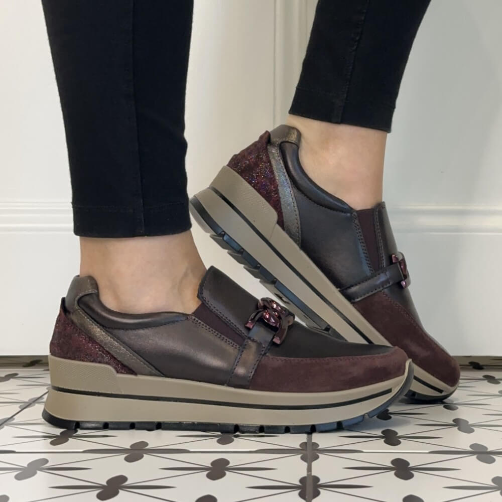 A person wearing Igi & Co   burgundy slip-on platform sneakers with a decorative buckle detail, paired with black pants, standing on a patterned tile floor.