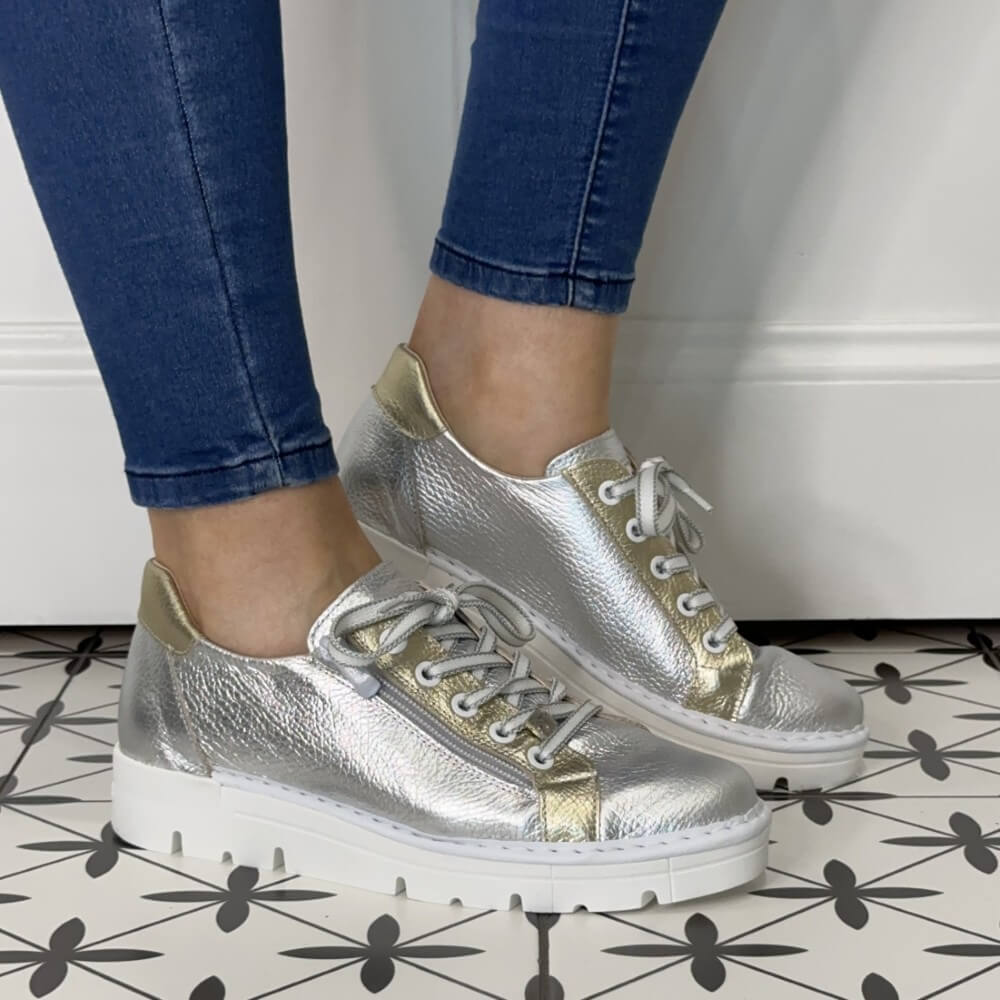 Silver and gold sneakers worn with blue jeans on a tiled floor.