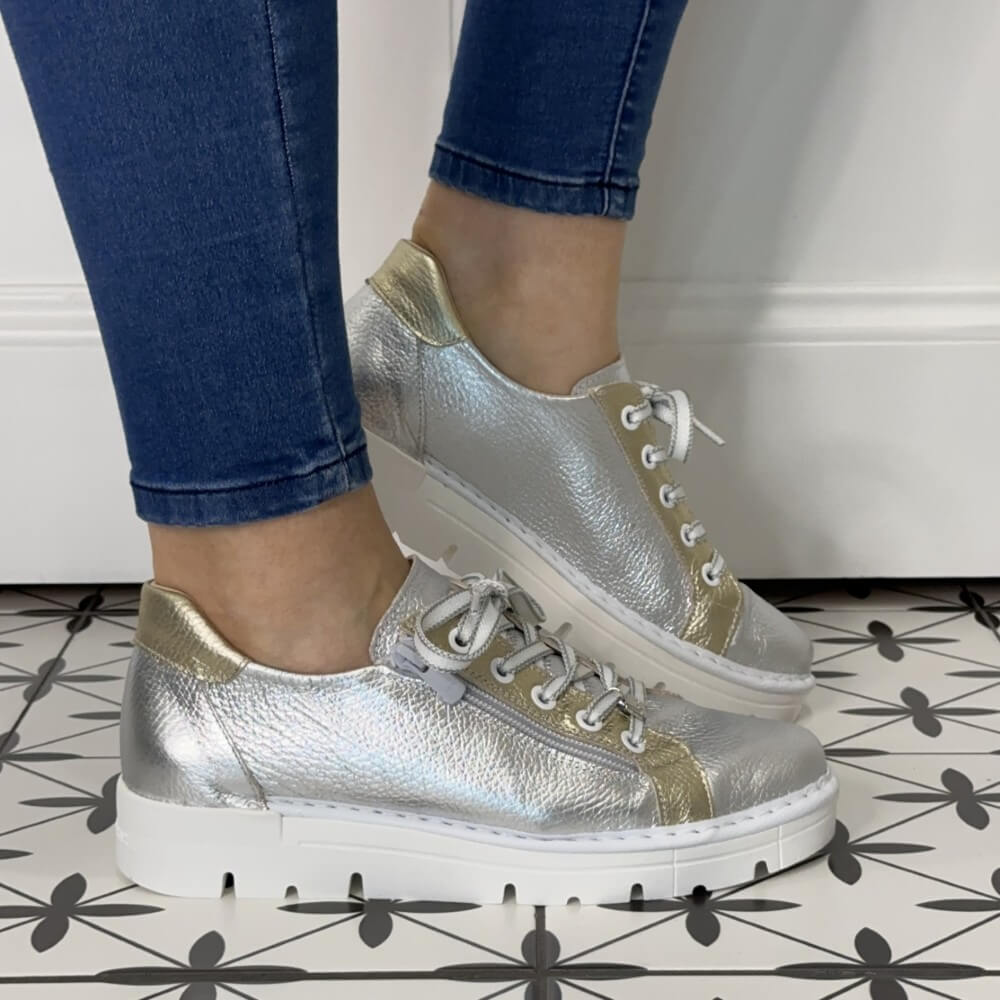 Silver and gold sneakers worn with blue jeans on a patterned floor.