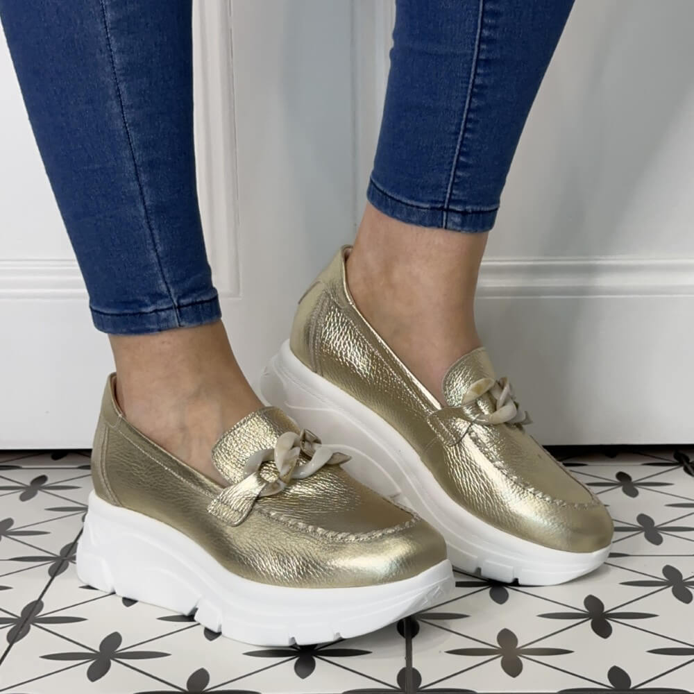 Gold loafers with white soles worn with blue jeans on a patterned floor.