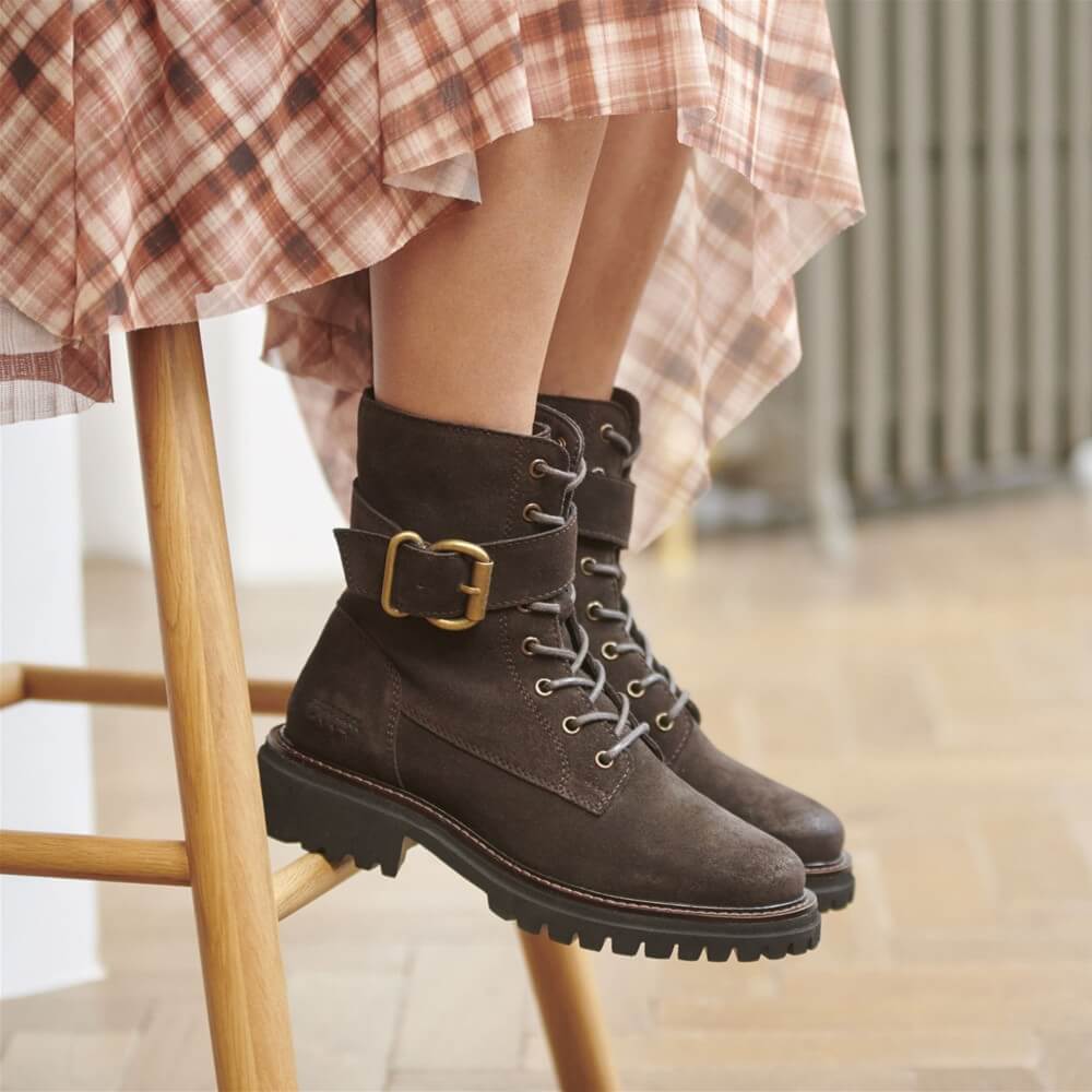 A person wearing Josef Seibel dark brown suede lace-up ankle boots with a wide strap and gold buckle, sitting on a wooden stool. The person is also wearing a brown and beige plaid skirt.