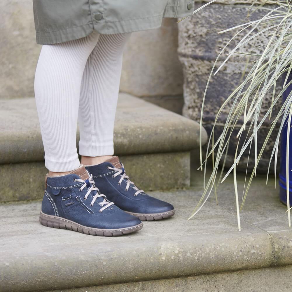 A Person wearing a pair of Josef Seibel navy blue leather ankle boots with white laces, paired with white ribbed tights and a khaki coat, standing on stone steps.