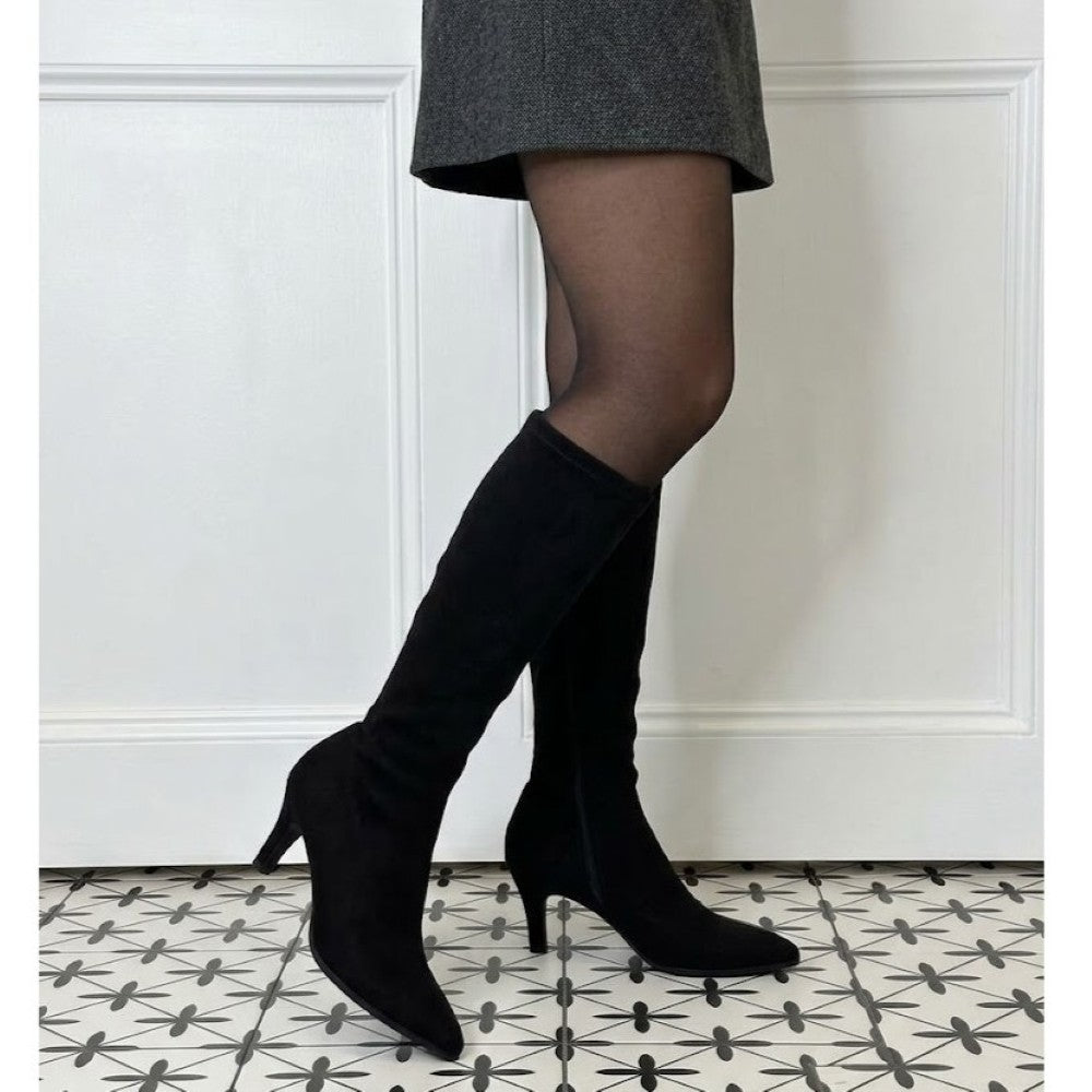 A person wearing Lodi black knee high heeled boots with pointed toes and mid-height stiletto heels, paired with sheer black tights and a grey skirt, standing on a black-and-white patterned tile floor against a white panelled wall.
