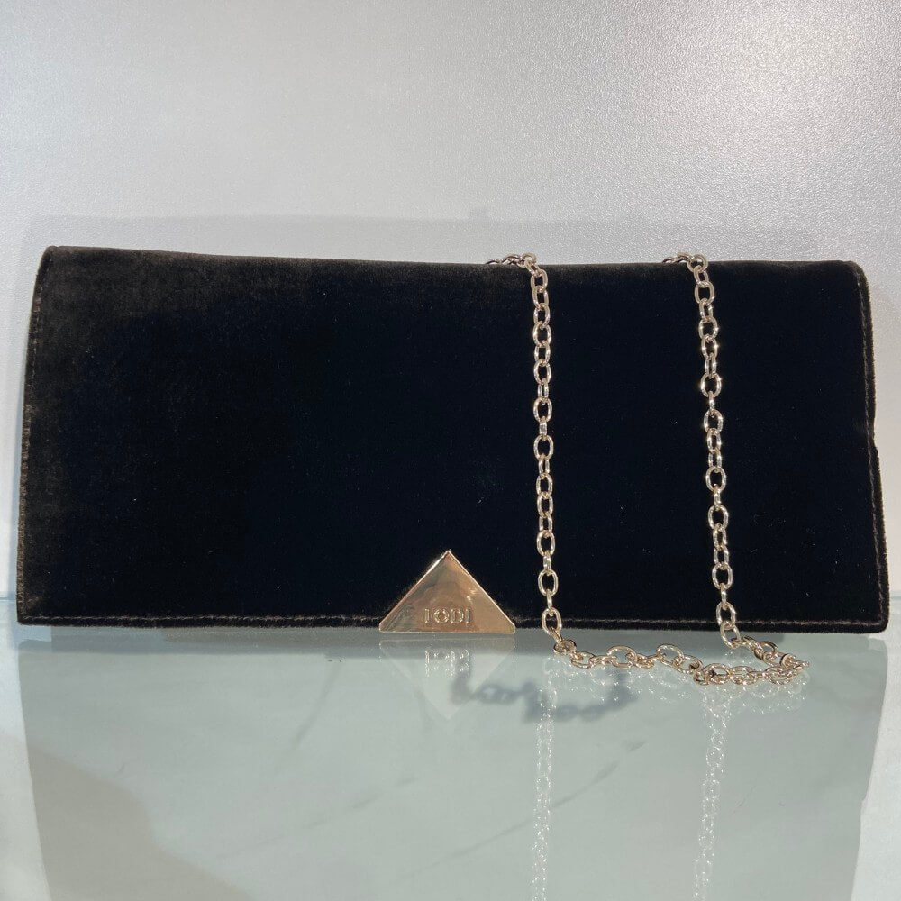 A Lodi Testa Brown velvet clutch bag with a gold-tone chain strap and a triangular gold-tone logo plaque on the front, displayed on a reflective light grey surface.