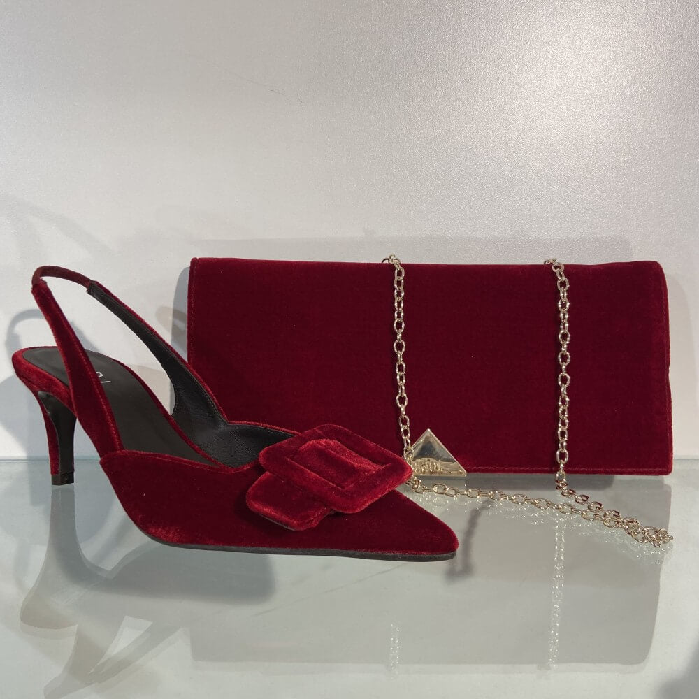 A Lodi red velvet pointed-toe slingback pump with a matching bow detail, displayed next to a matching red velvet clutch bag with a gold chain strap on a reflective white surface.