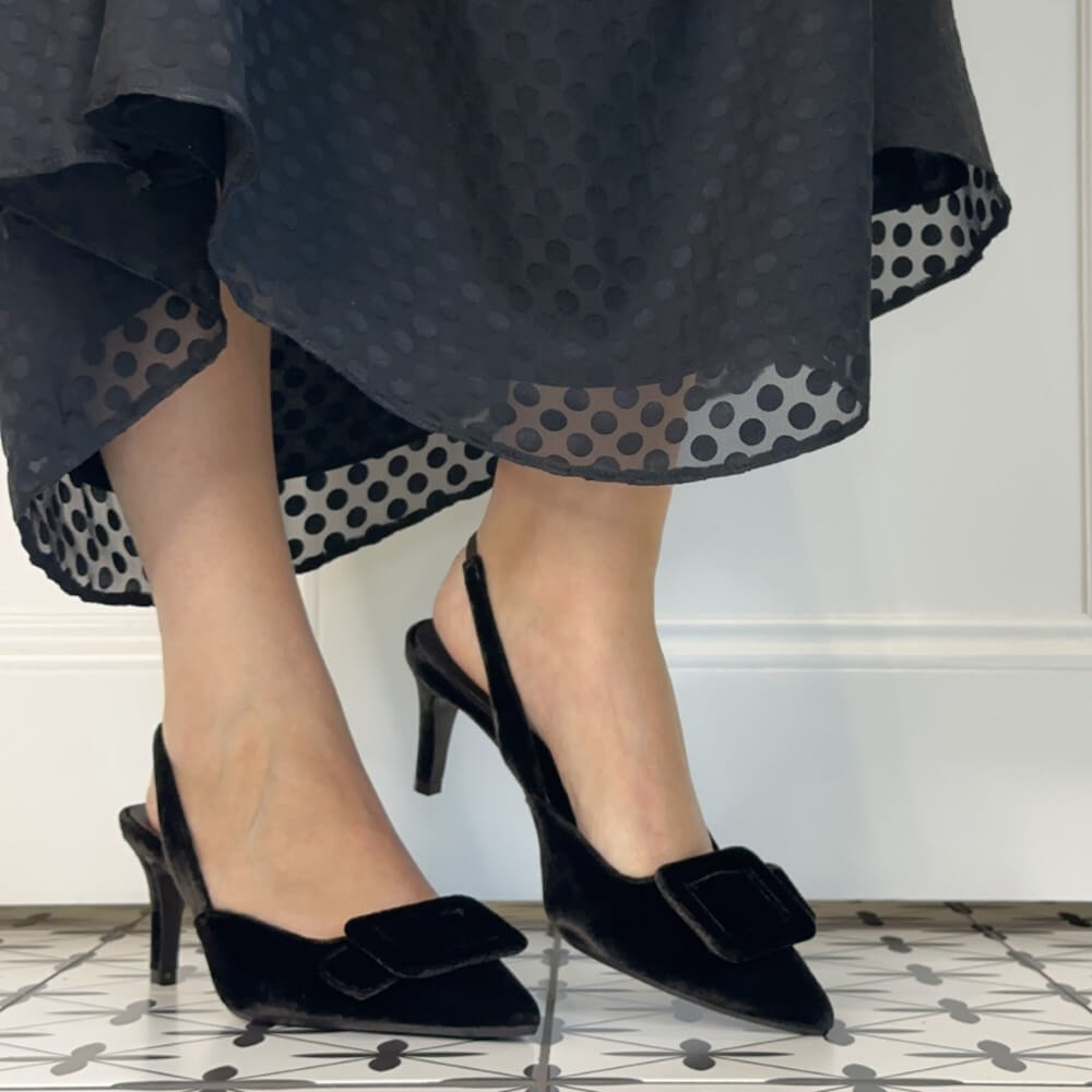 Lodi Testa Brown velvet slingback kitten heel pumps with bow details, shown on a model wearing black patterned dress against a white wall and tiled floor.